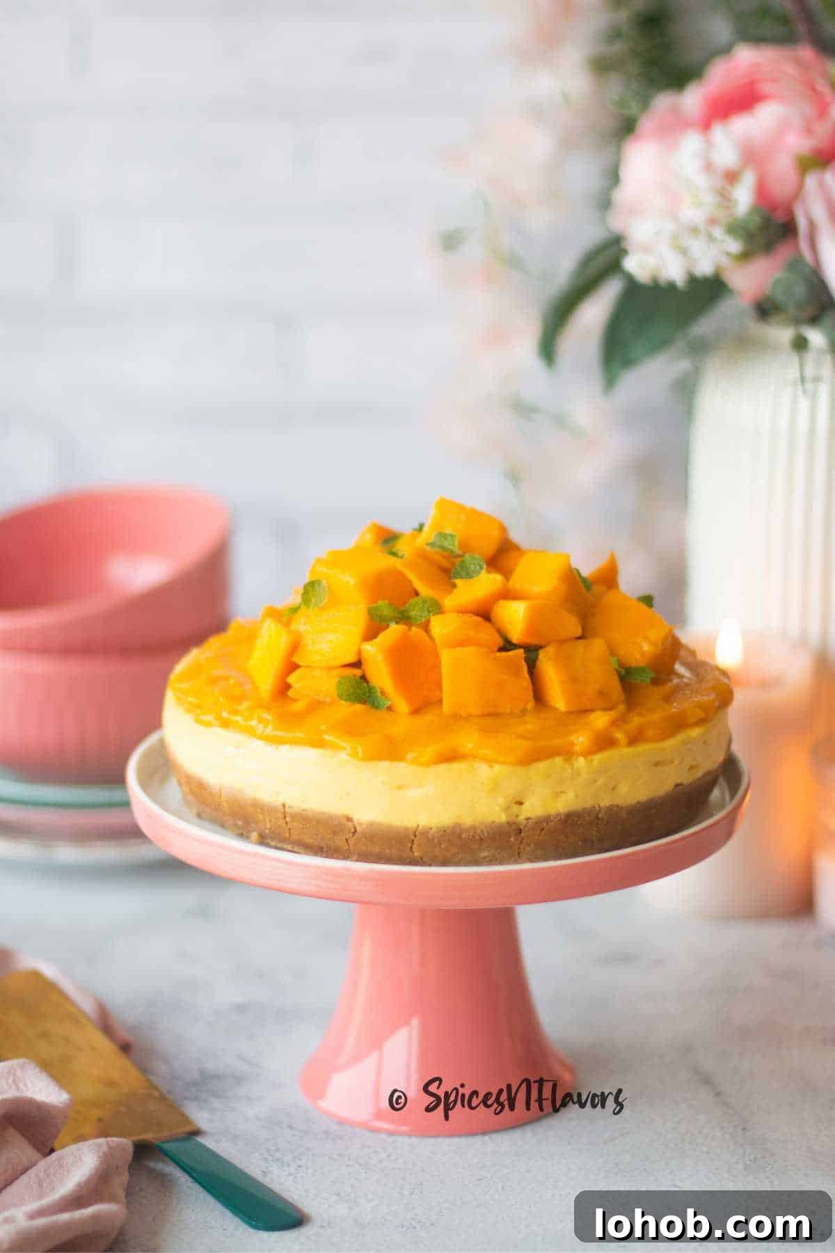 Eggless Baked Mango Cheesecake Recipe: Creamy Tropical Slice 2 baked version of mango cheesecake garnished with fresh mango pulp, mango pieces and mint leaves is placed on a pink cake stand with flowers and candle in the background
