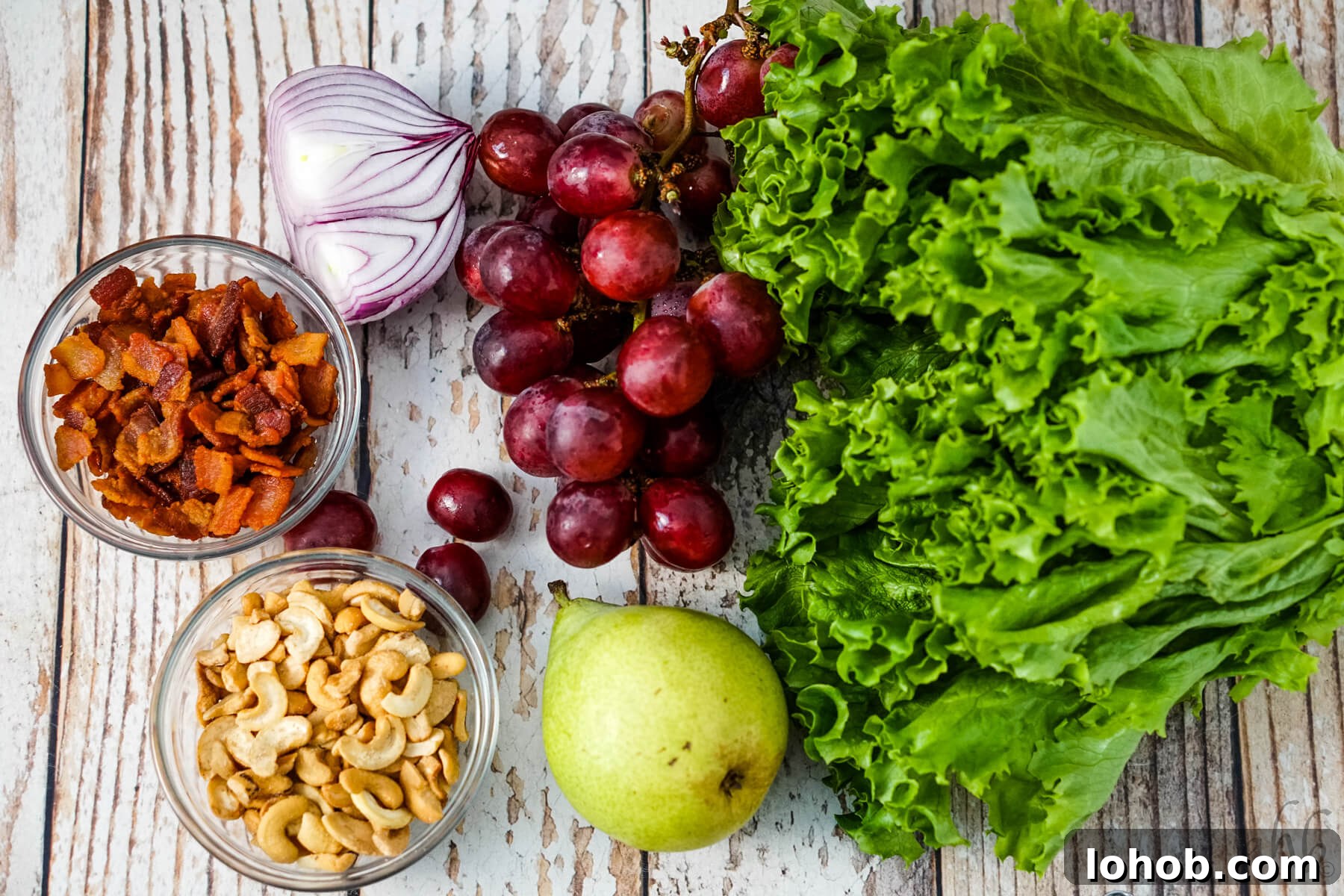 Crunchy Cashew Salad with Lime-Ginger Dressing 5 A head of lettuce, a pear, some red seedless grapes, half of a red onion, a bowl of bacon pieces and a bowl of cashew halves and pieces sit next to each other.