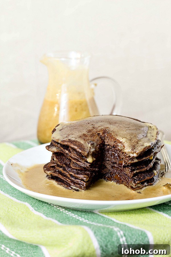Yeasted Chocolate Chip Pancakes with Rich Cocoa Batter 2 A tall stack of chocolate pancakes with chocolate chips on a white plate with syrup poured over them and a pitcher of coffee maple syrup in the background.