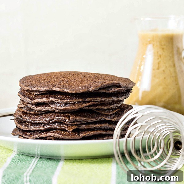 Yeasted Chocolate Chip Pancakes with Rich Cocoa Batter 3 A plate of stacked chocolate yeast raised pancakes on a white plate with a whisk and a pitcher of coffee maple syrup in the background.