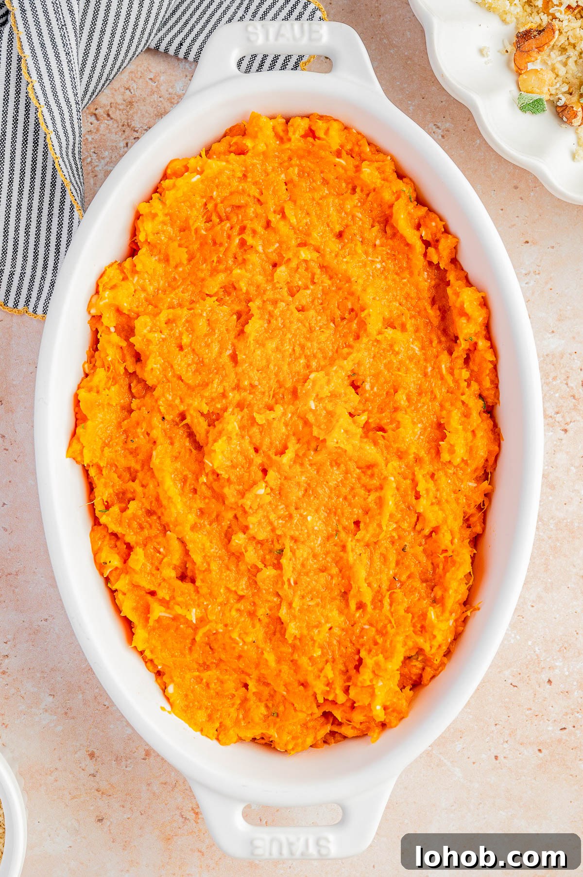 Oval white dish filled with mashed sweet potatoes on a light countertop, with part of a cloth napkin and another dish partially visible nearby.