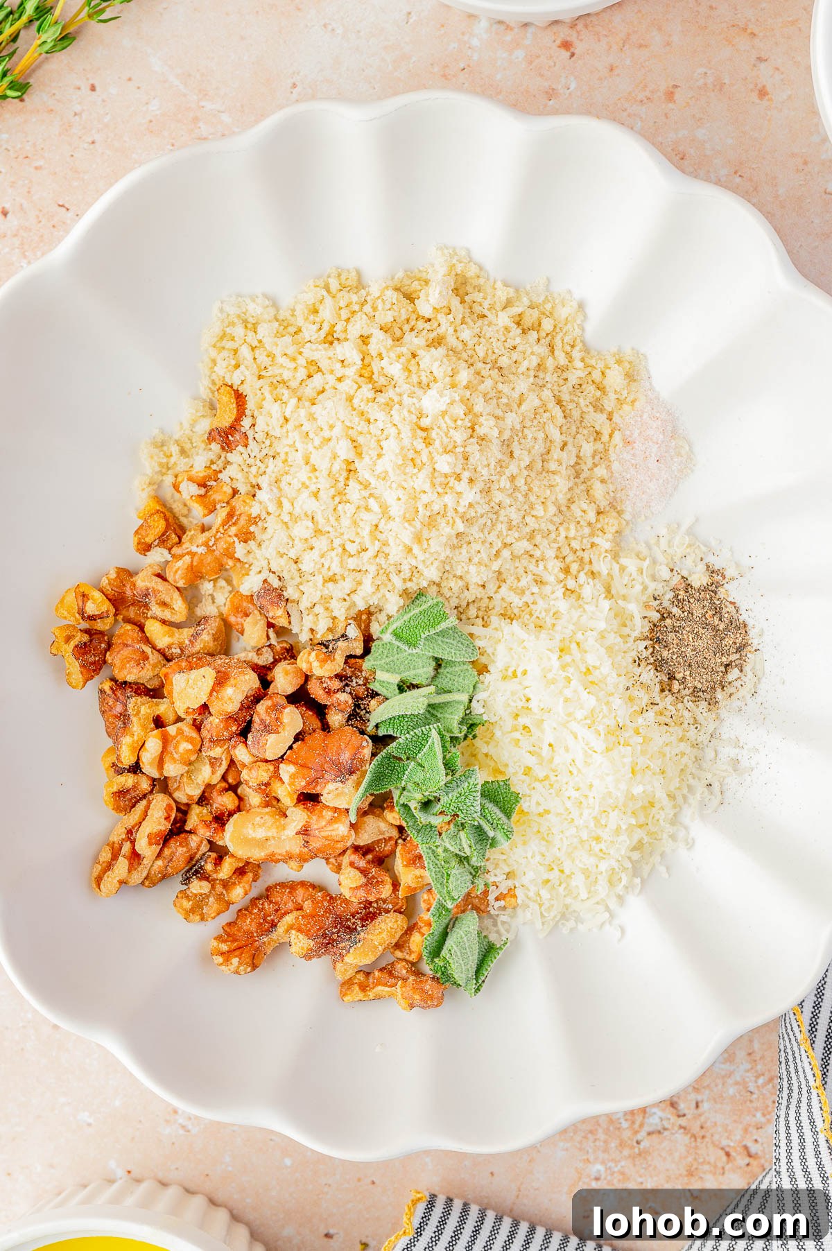 White plate with piles of panko breadcrumbs, chopped walnuts, grated cheese, black pepper, fresh sage leaves, and salt on a light countertop.