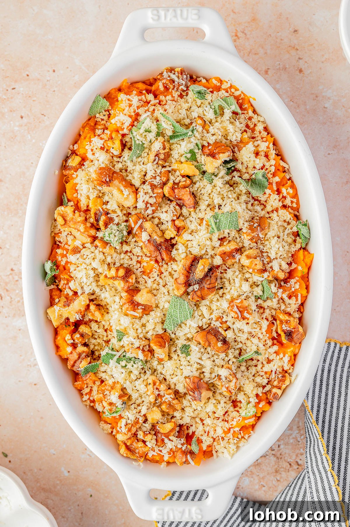 A white oval baking dish filled with a baked casserole topped with breadcrumbs, chopped nuts, and fresh herbs on a light surface.
