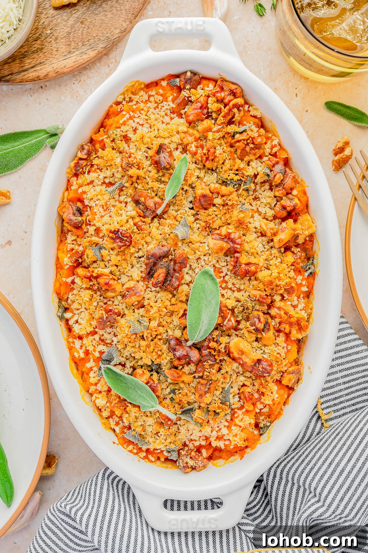 A baked casserole topped with breadcrumbs, walnuts, and sage leaves is presented in a white oval dish.