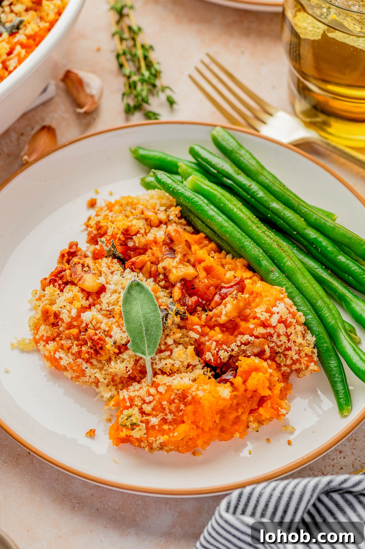 A plate with a serving of baked sweet potato casserole topped with breadcrumbs, garnished with a sage leaf, and green beans on the side.