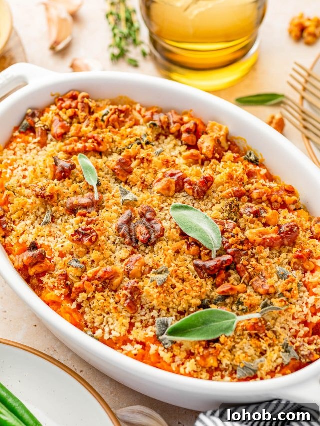 A baked casserole topped with golden breadcrumbs, chopped nuts, and fresh sage leaves, served in a white oval dish on a table.
