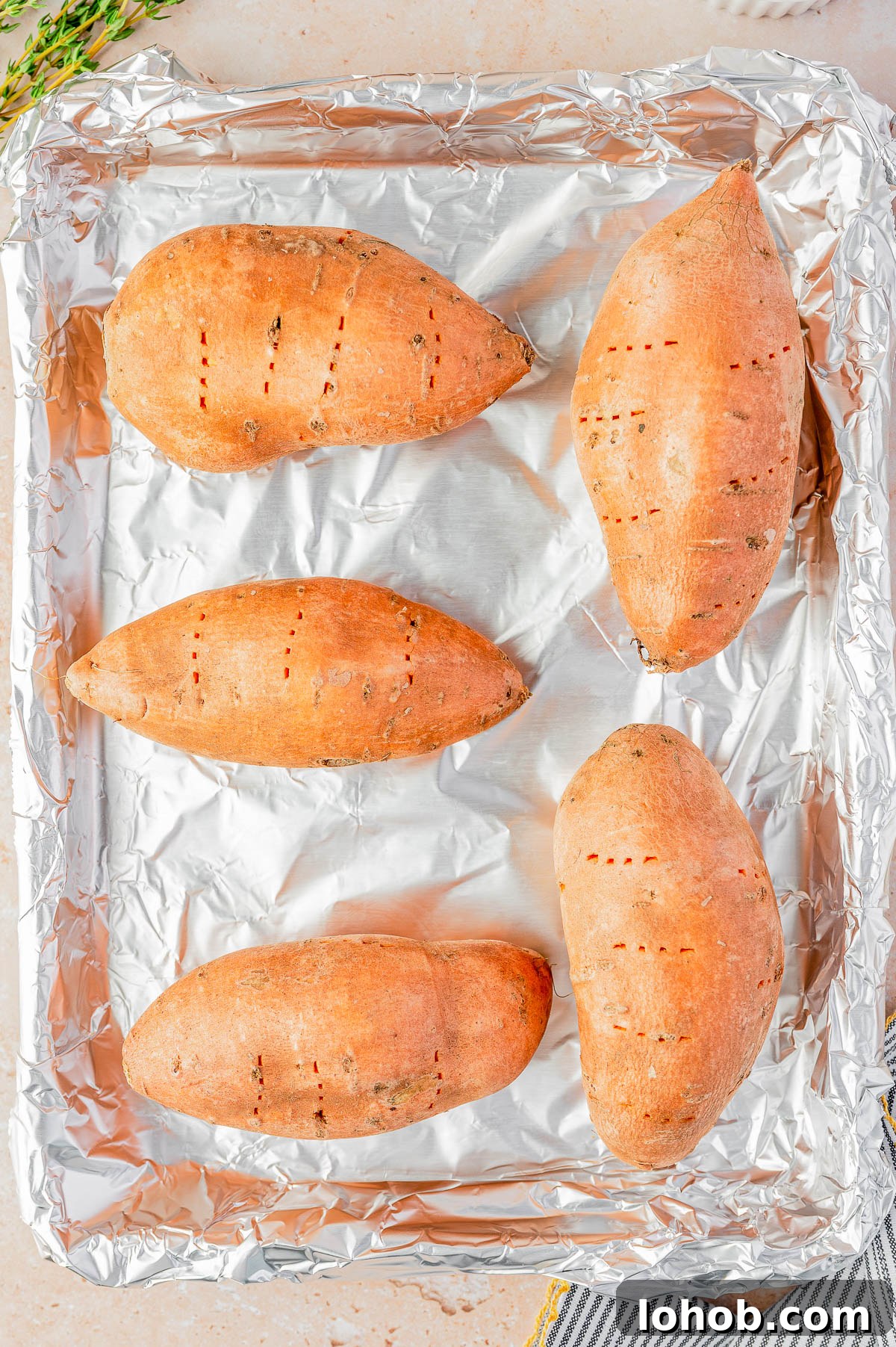 Four whole sweet potatoes with fork holes are placed on a foil-lined baking sheet, ready for roasting.