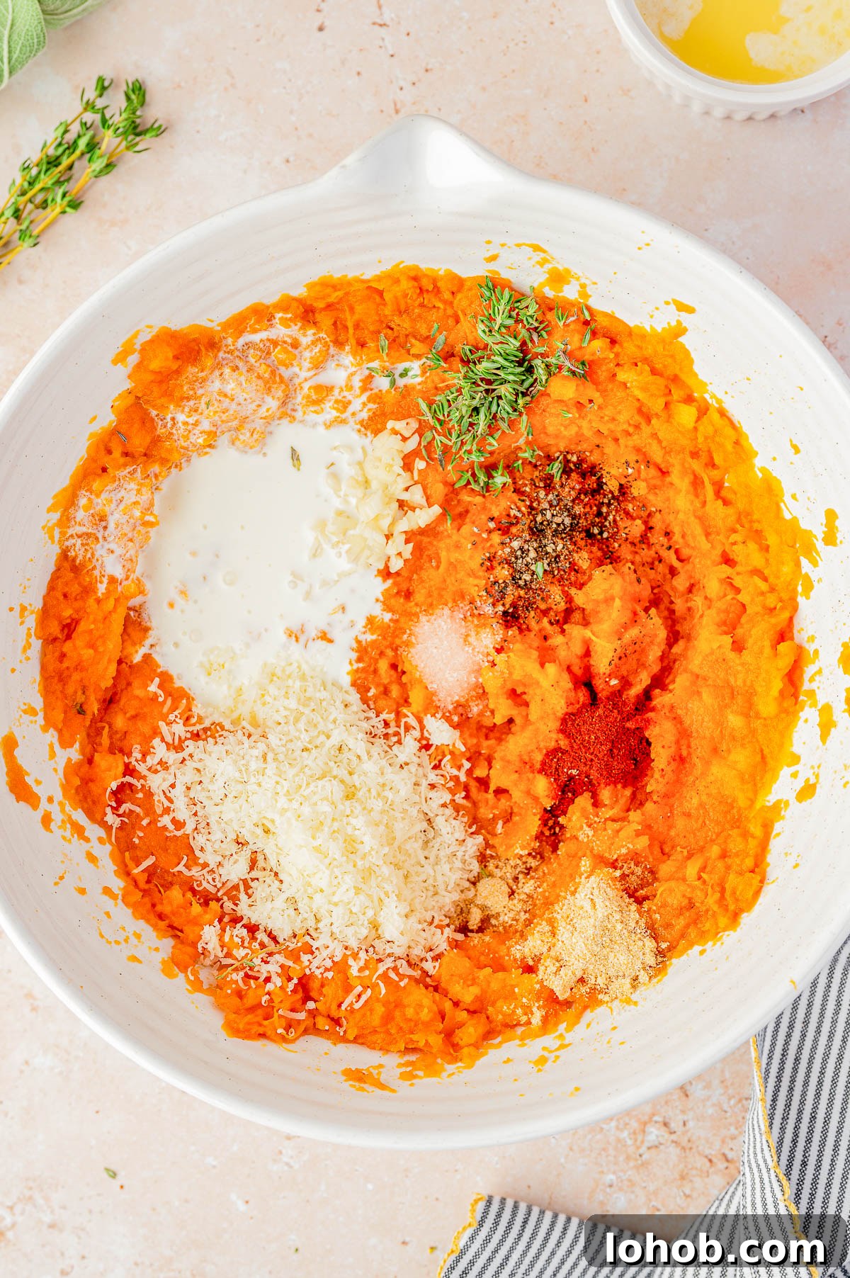A bowl of mashed sweet potatoes with added cream, grated cheese, spices, minced garlic, salt, pepper, and fresh thyme on top, ready to be mixed.