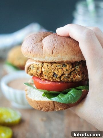 Close up shot of a hand holding a veggie burger on a wheat bun with spinach leaves, a tomato slice, and bbq sauce.