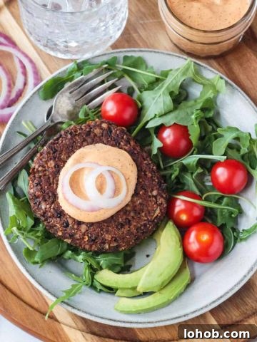 Overhead view of a veggie burger topped with sauce and onions on a bed of lettuce with tomatoes and avocado next to it.