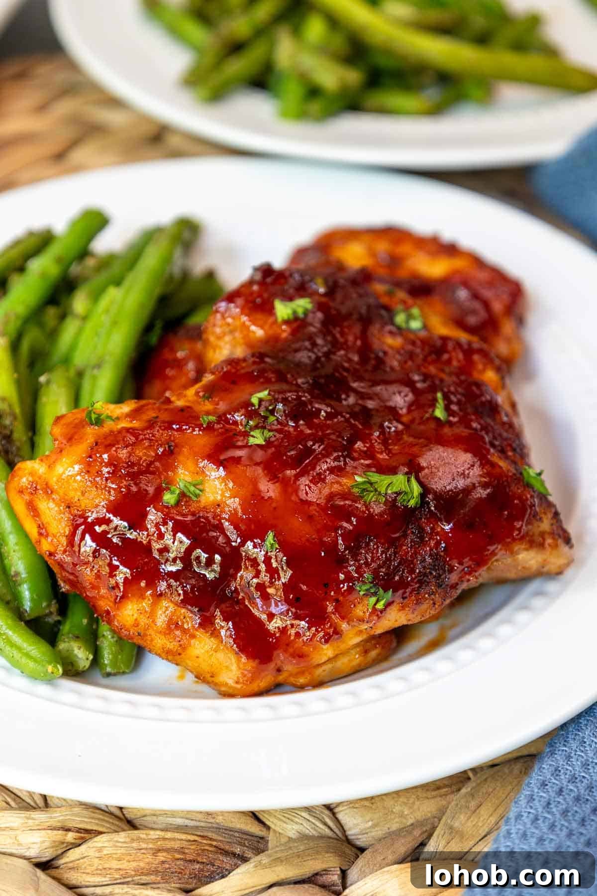 Crispy Oven-Baked BBQ Boneless Chicken Thighs 2 Close-up of oven baked BBQ boneless chicken thighs glazed with sauce and sprinkled with parsley, served with green beans on a white plate.