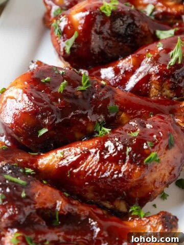 A close view of several barbecue glazed chicken drumsticks shows the thick, shiny coating and small herb garnish on top. The texture of the sauce and crisped skin is clearly visible.