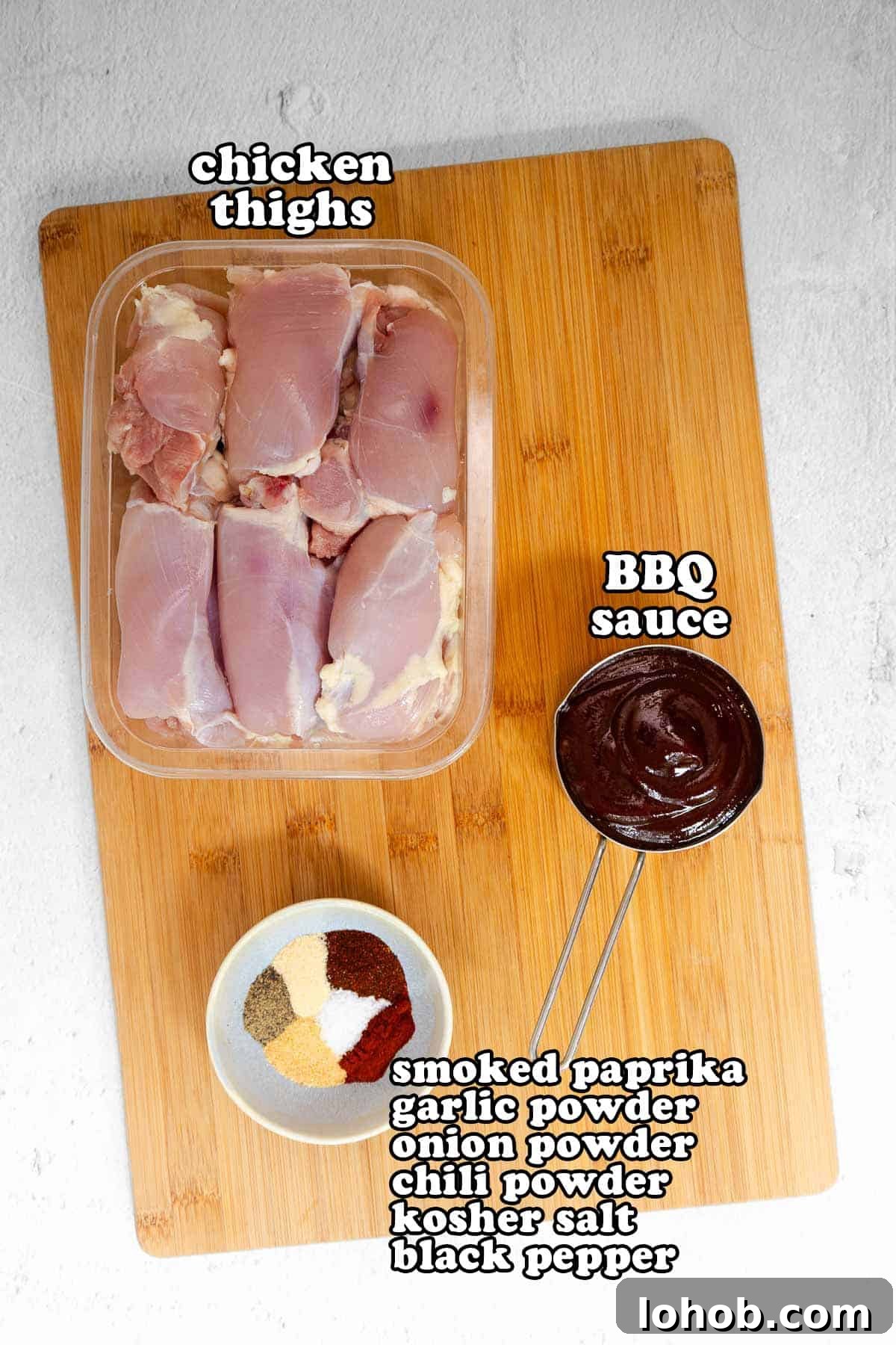 Crispy Oven-Baked BBQ Boneless Chicken Thighs 3 Flat lay of raw ingredients on a wooden cutting board including boneless chicken thighs, a bowl of BBQ sauce, and a spice blend labeled with smoked paprika, garlic powder, onion powder, chili powder, kosher salt, and black pepper.