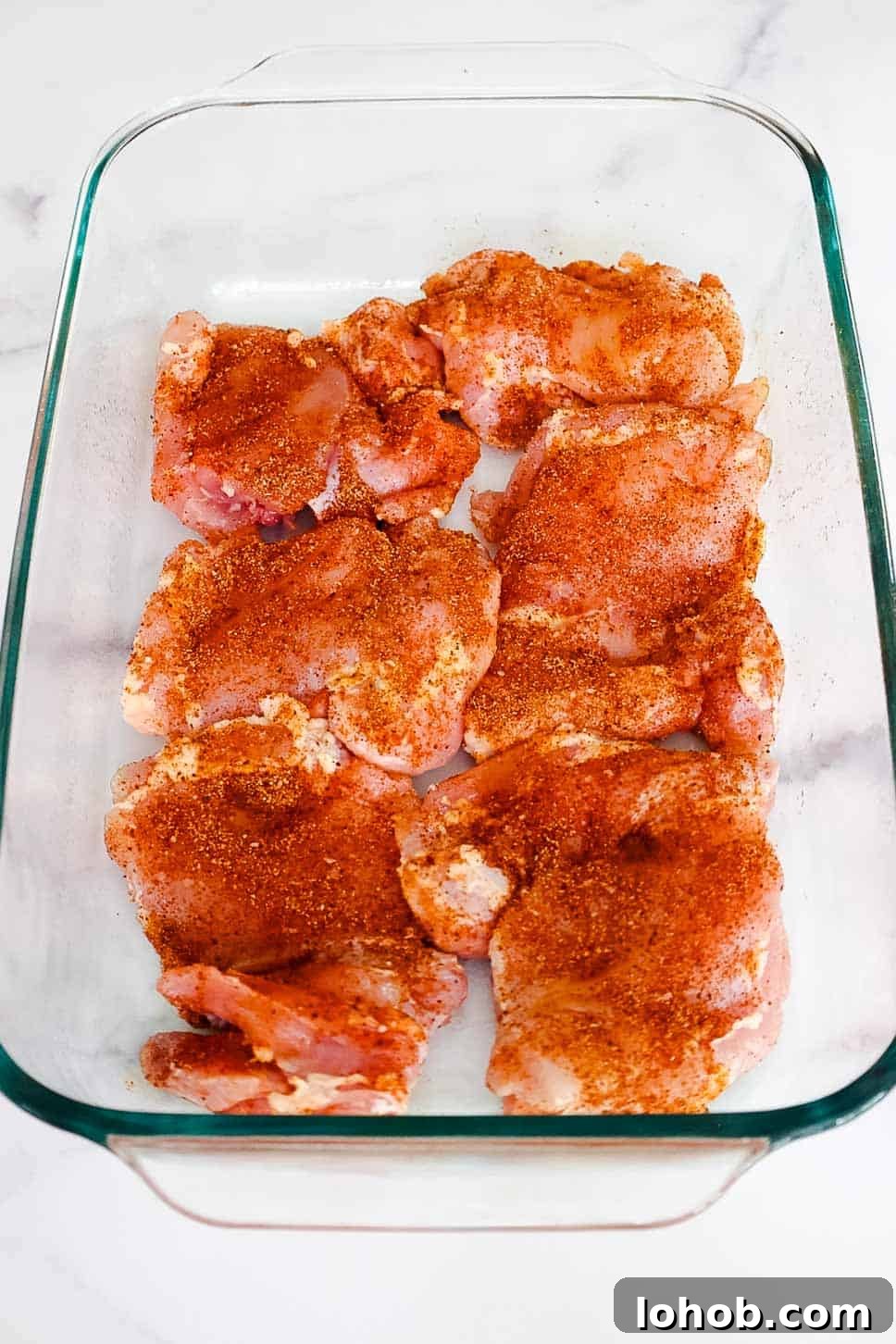 Crispy Oven-Baked BBQ Boneless Chicken Thighs 4 Raw boneless chicken thighs evenly coated in dry spice rub, arranged in a glass baking dish before cooking.