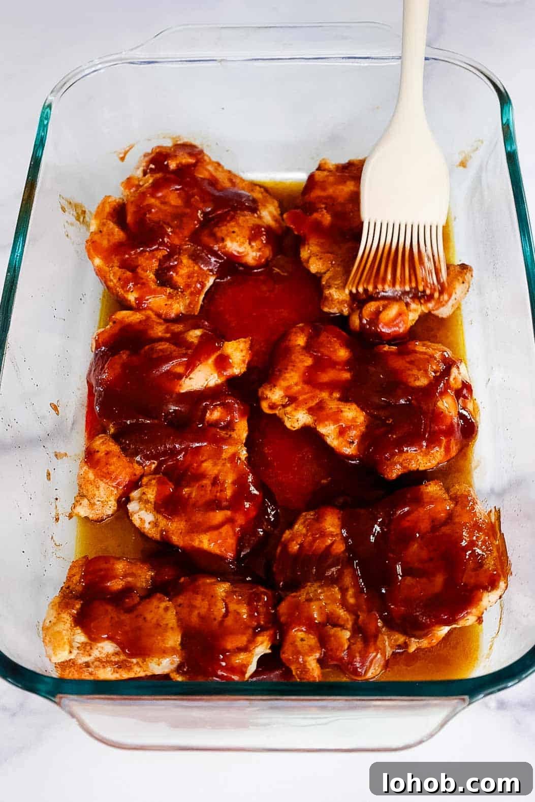 Crispy Oven-Baked BBQ Boneless Chicken Thighs 6 Chicken thighs being brushed with BBQ sauce using a silicone brush, midway through baking in a glass dish.
