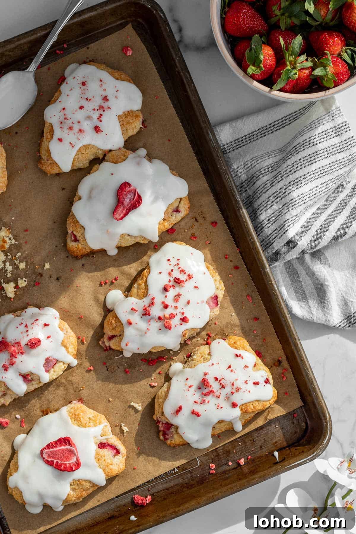 Strawberry Cream Scones with Vanilla Glaze 2 strawberry and cream scones with a vanilla glaze on a baking sheet with a bowl of strawberries next to it