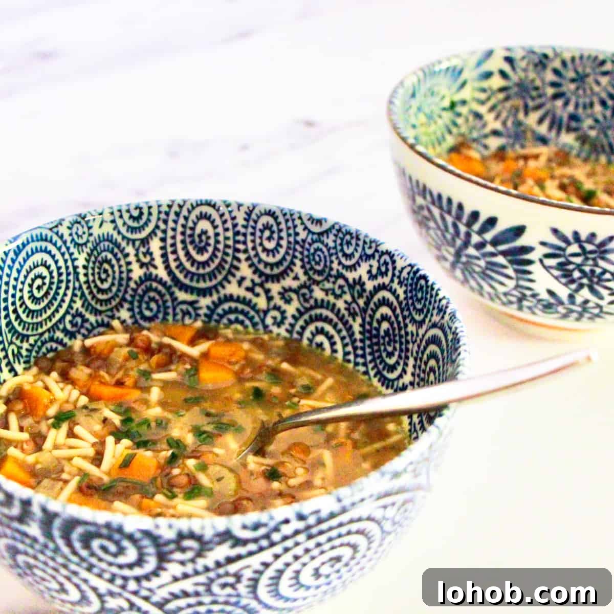 Hearty Vegan Lentil and Vegetable Noodle Soup Recipe 2 Two blue and white decorated bowls containing veggie noodle soup, spaced with one in front of the other, on a white and grey marble surface.