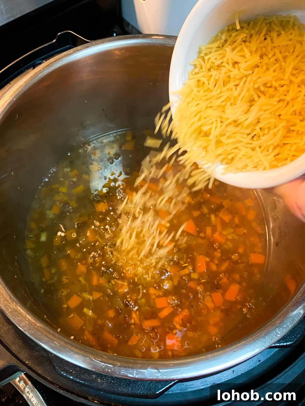 Hearty Vegan Lentil and Vegetable Noodle Soup Recipe 9 Photo showing cooked soup with noodles being added from a bowl.