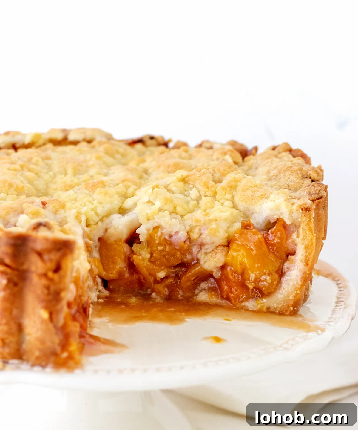 Perfect Peach Pie with Buttery Crumb Topping 3 Peach crumble pie on a white cake stand, piece missing. White background.