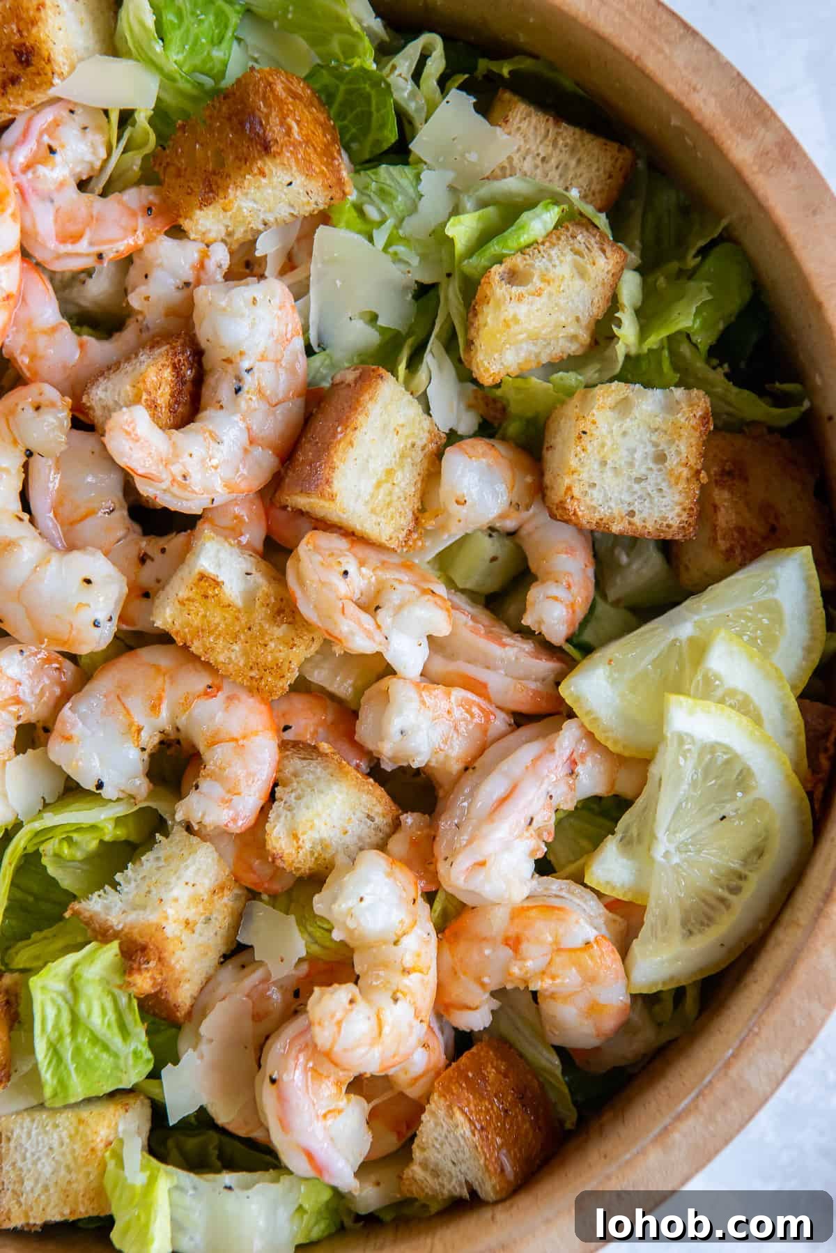 Grilled Shrimp Caesar Salad with Crispy Parmesan Croutons 12 A top down close up of roasted shrimp on a bed of romaine lettuce with croutons and slices of lemon in a wood bowl.