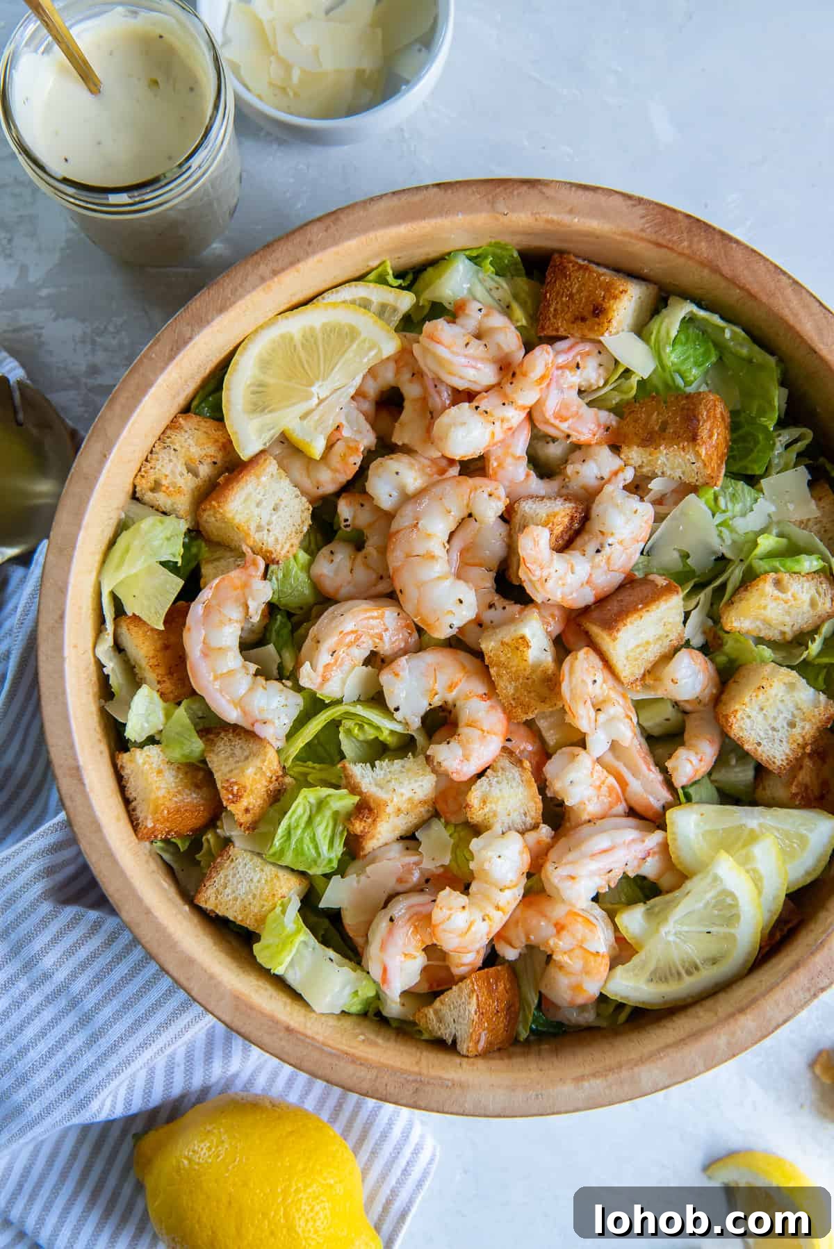 Grilled Shrimp Caesar Salad with Crispy Parmesan Croutons 3 A top down shot of Shrimp Caesar Salad in a wood bowl near a mason jar filled with dressing and fresh lemons.