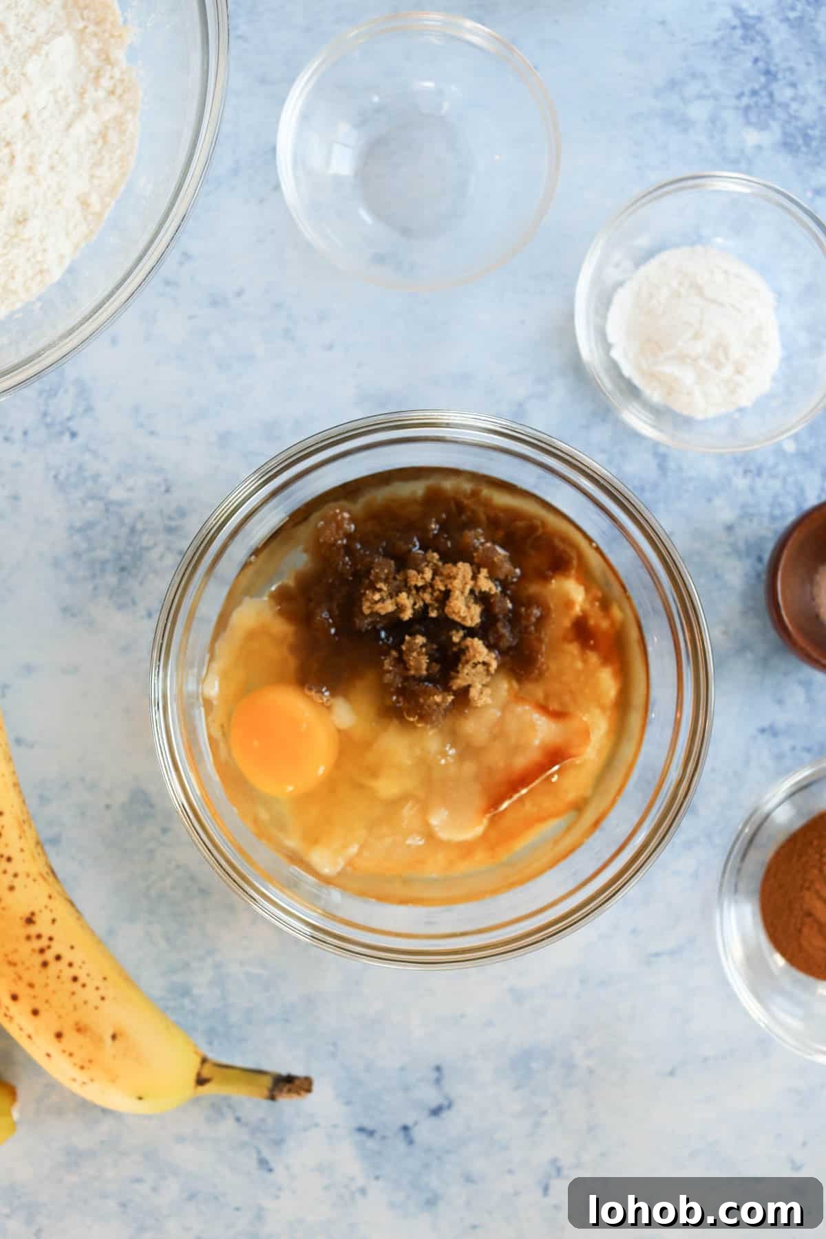 A bowl of bananas, eggs, flour and other ingredients on a table.