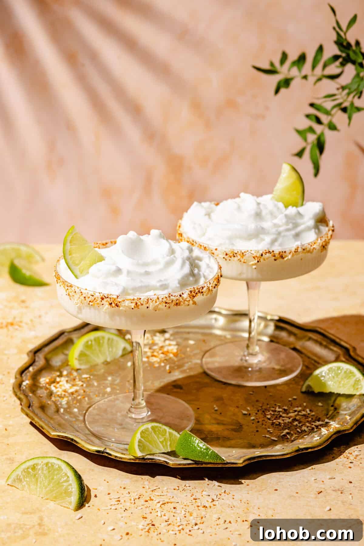 Coconut Frozen Margarita Recipe — Creamy Tropical Cocktail 2 Two Frozen Coconut Margaritas in coupe cocktail glasses, each garnished with a lime wedge. The margaritas sit on a bronze tray on top of a tan table, with lime wedges on both the tray and the table. A green branch is visible in the top right corrner.
