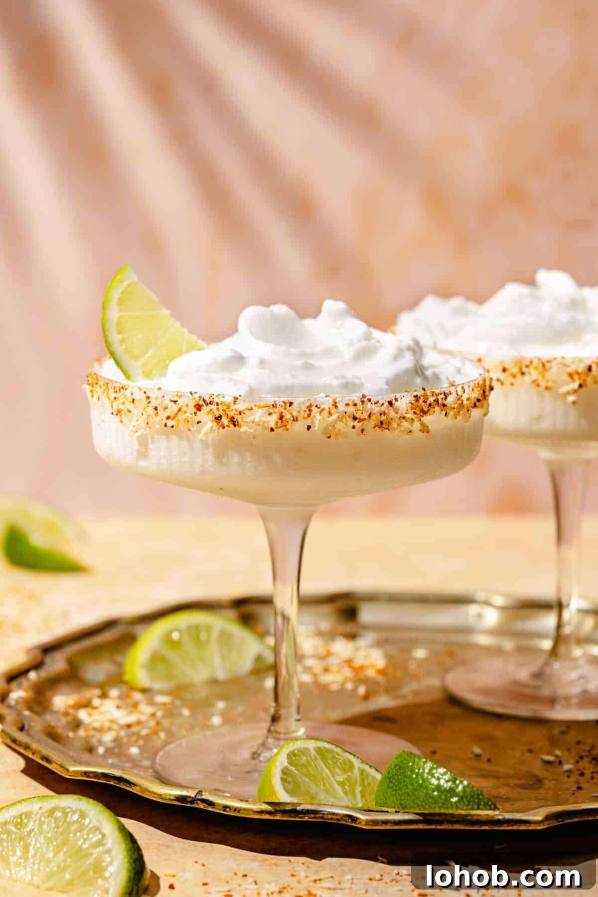 Coconut Frozen Margarita Recipe — Creamy Tropical Cocktail 4 Straight shot of two coupe cocktail glasses, each containing a frozen coconut margarita. The glass in the forefront is garnished with a lime wedge, and there are limes on both the bronze tray the glasses are on, as well as on the table.