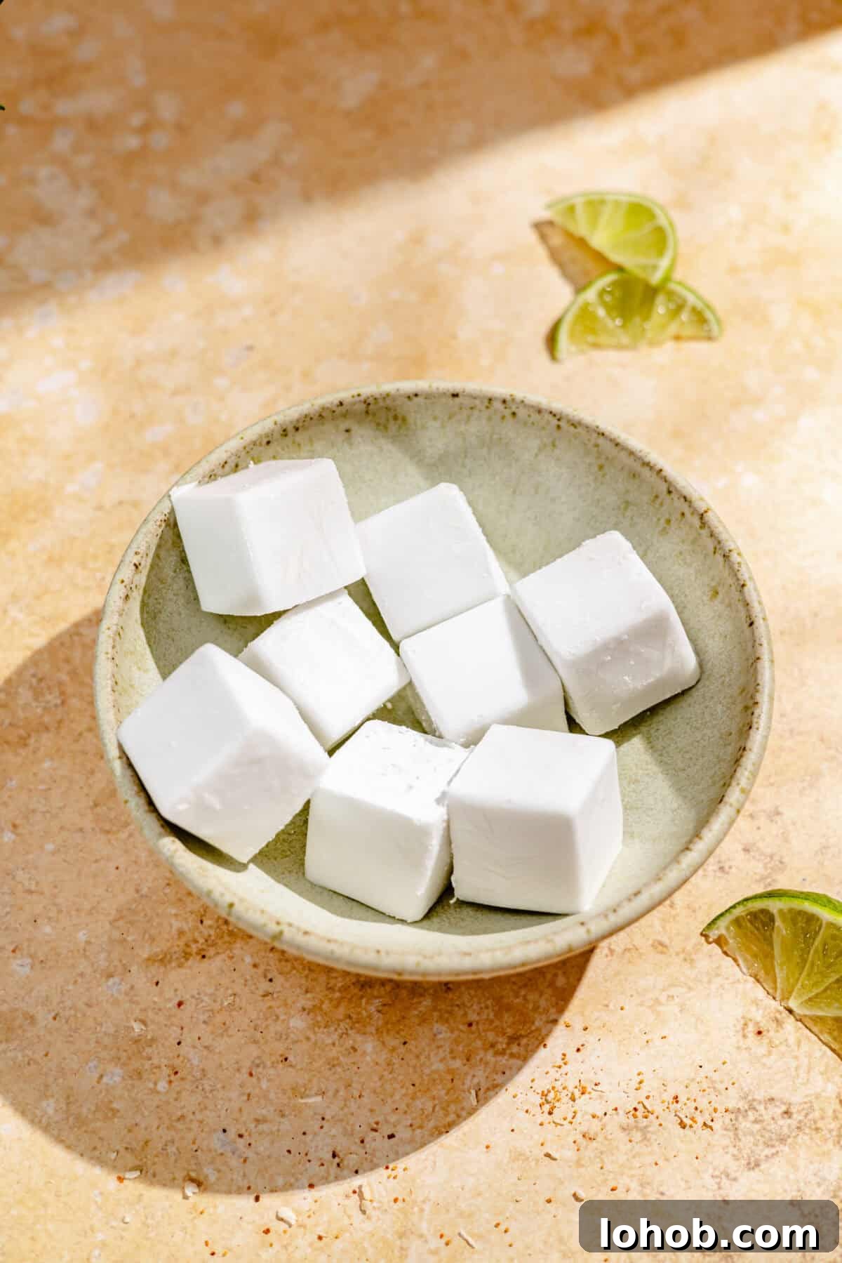 Coconut Frozen Margarita Recipe — Creamy Tropical Cocktail 5 A beige bowl with cubes of frozen coconut milk. Three lime wedges are visible, and the bowl rests on a tan table.