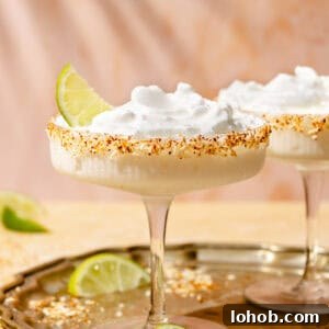 Straight shot of two coupe cocktail glasses, each containing a frozen coconut margarita. The glass in the forefront is garnished with a lime wedge, and there are limes on both the bronze tray the glasses are on, as well as on the table.