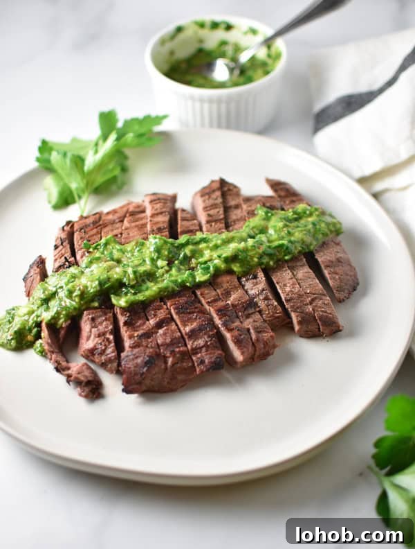 Grilled Flank Steak with zesty Chimichurri Sauce 3 Sliced grilled steak with herb chimichurri on top on a marble table with a bowl of chimichurri in the background