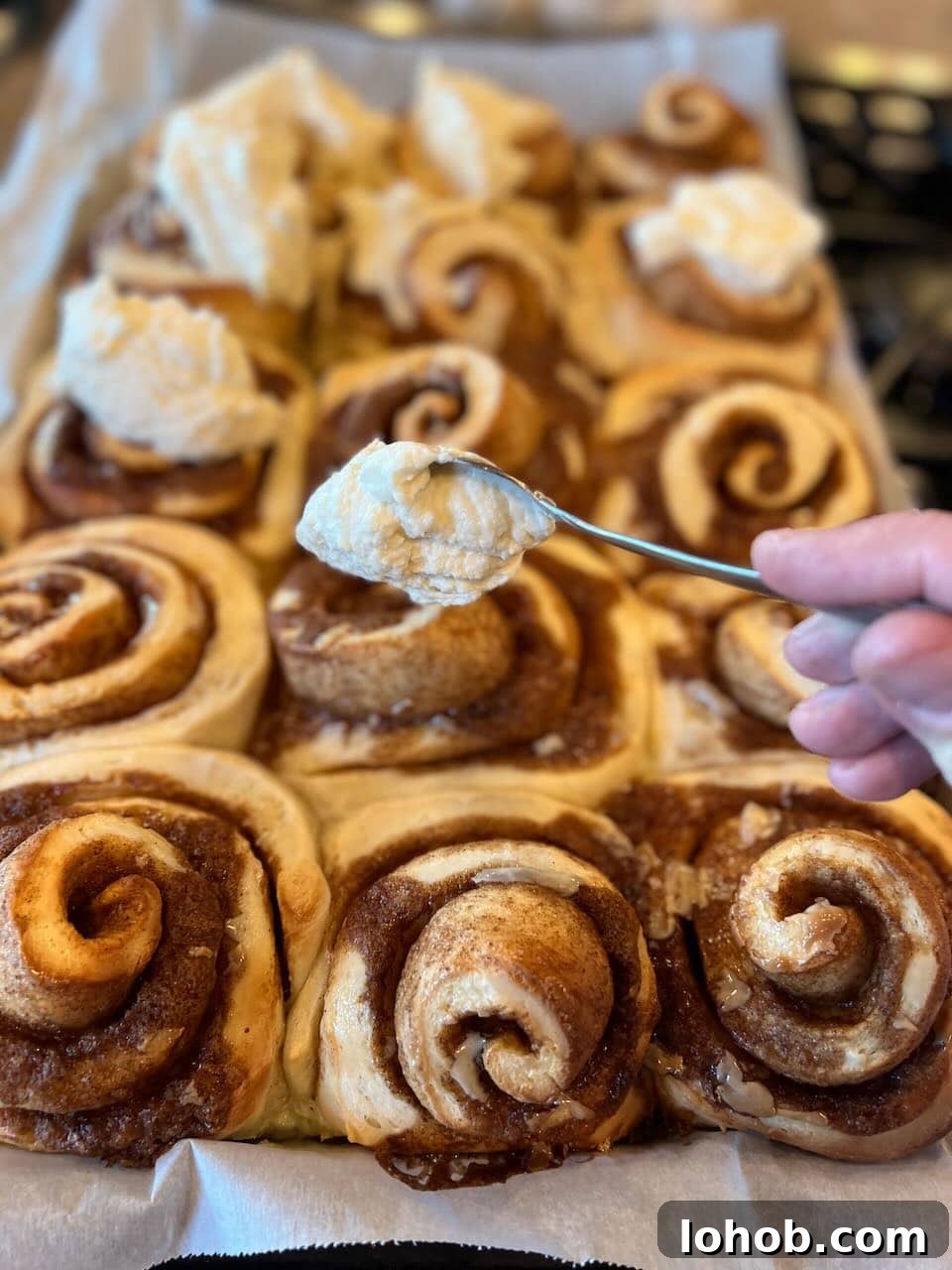 Soft Gooey Cinnamon Rolls Recipe for Perfectly Fluffy Layers 16 A hand dollops frosting with a spoon onto large cinnamon rolls.