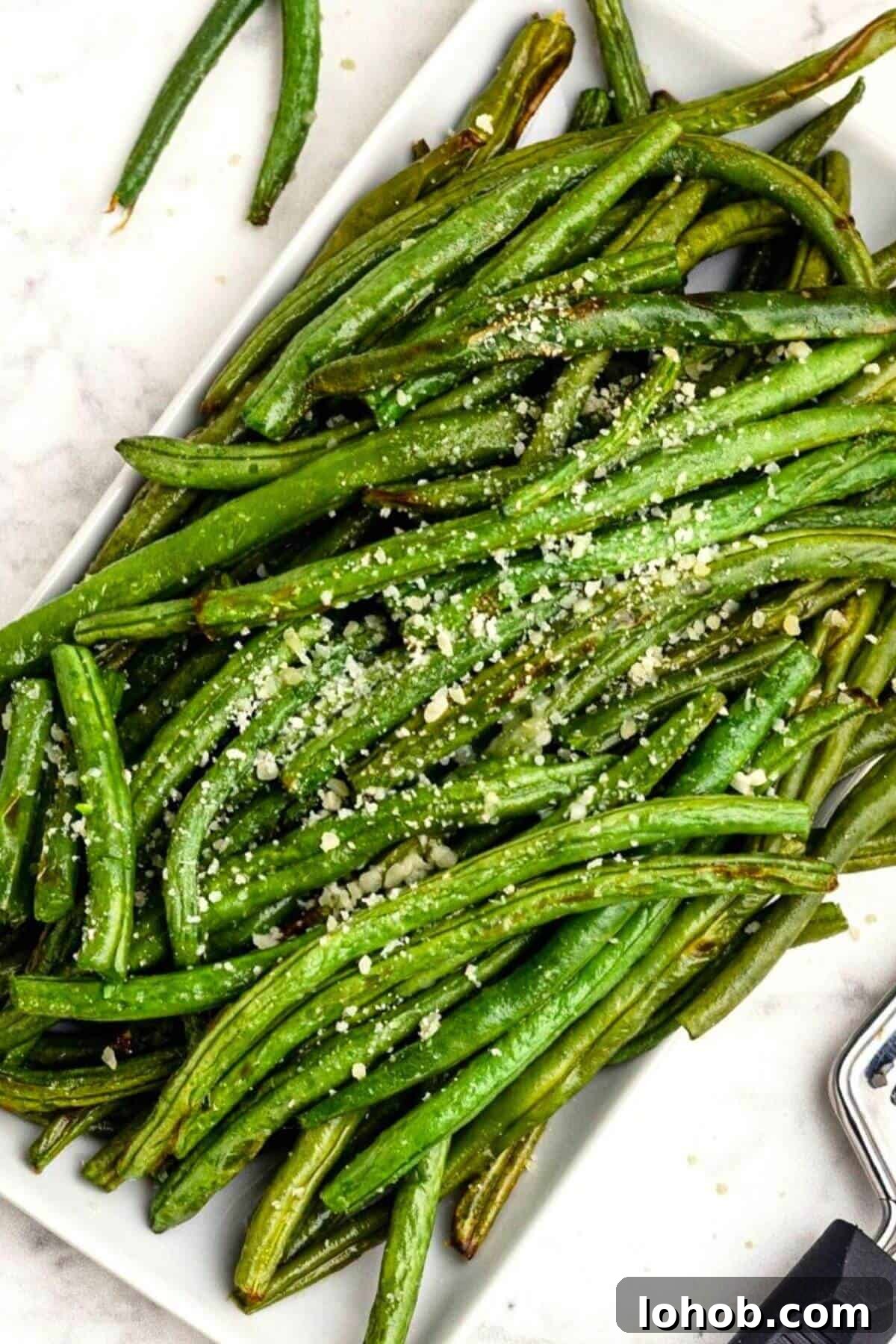 Crispy Air Fryer Green Beans with Garlic and Parmesan 2 Green juicy green beans stacked on a white rectangle plate seasoned and sprinkled with grated parmesan cheese.