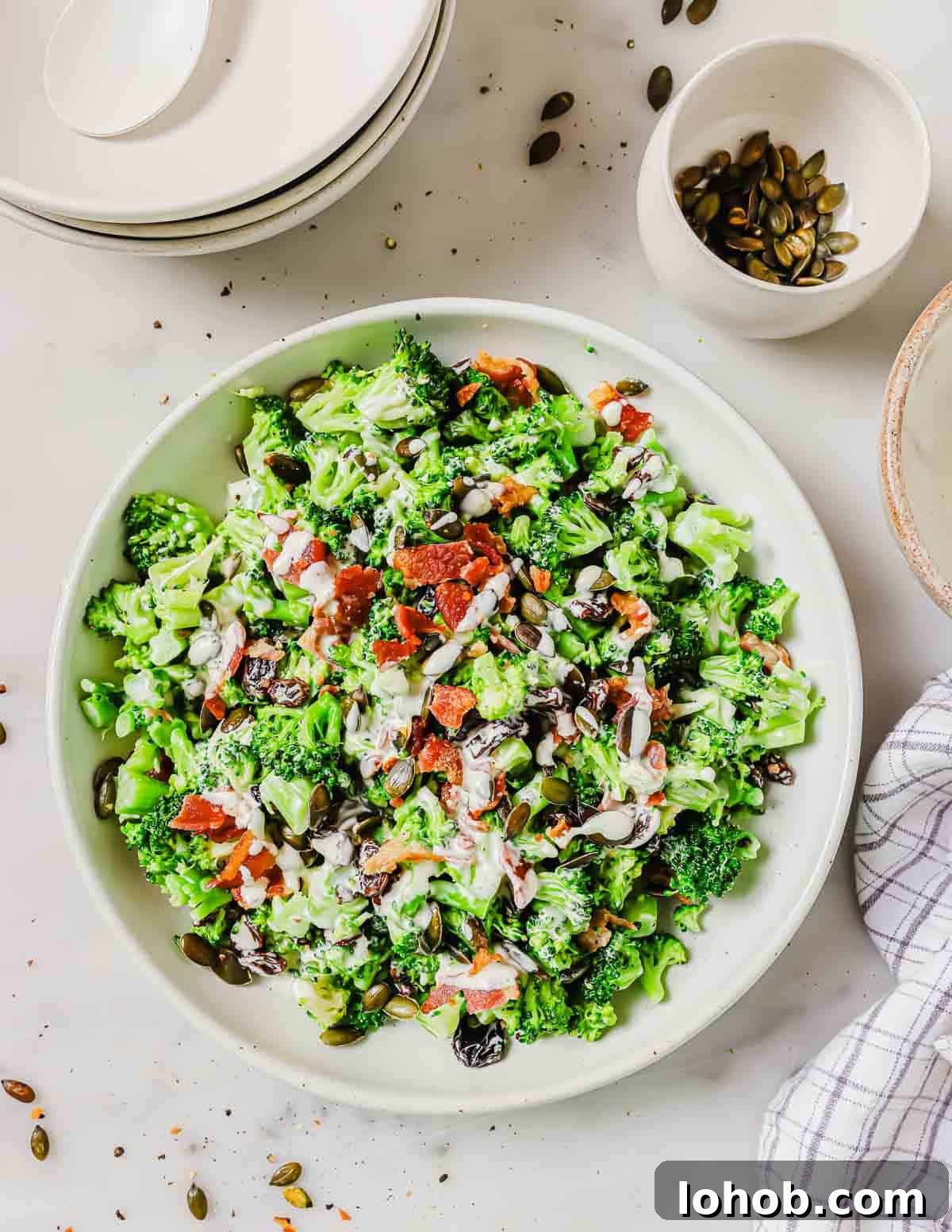 A bowl of broccoli salad with bacon pieces, and raisins sits next to a stack of plates and a small bowl of pumpkin seeds.
