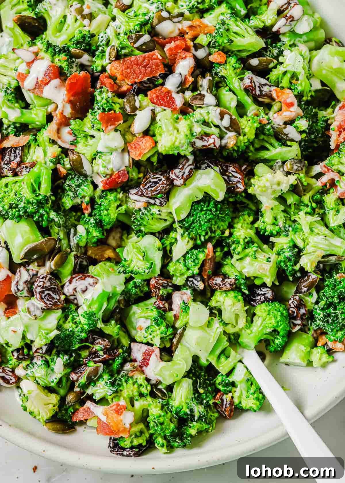 A close-up of a broccoli raisin salad mixed with bacon bits and seeds served on a white plate with a white utensil visible to the side.
