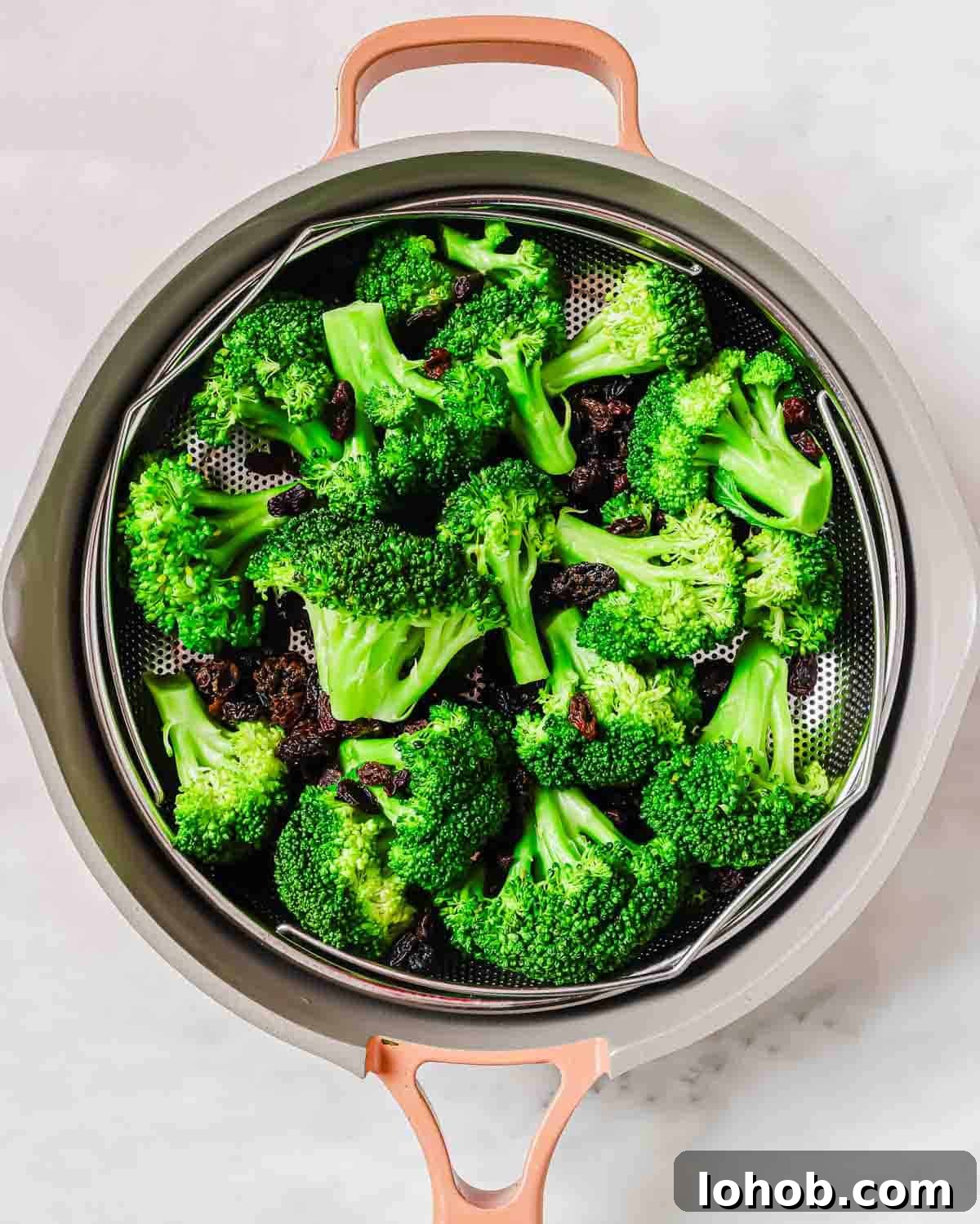Steamed broccoli florets in a metal colander inside a beige pot with copper handles on a light countertop.