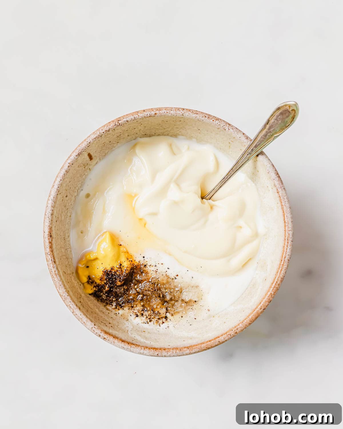 A bowl containing dressing ingredients including mayonnaise, mustard, ground black pepper, and diced garlic cloves with a spoon.