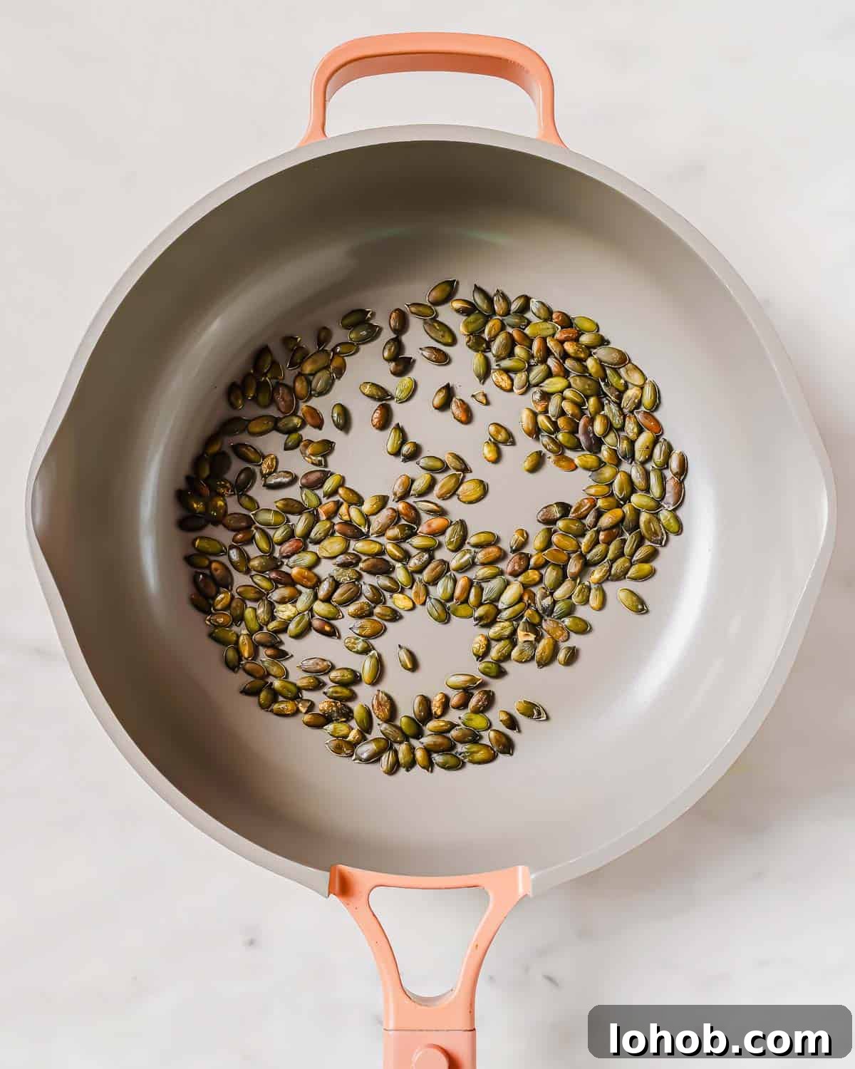 A gray frying pan with a pink handle containing a single layer of roasted pumpkin seeds on a white marble surface.