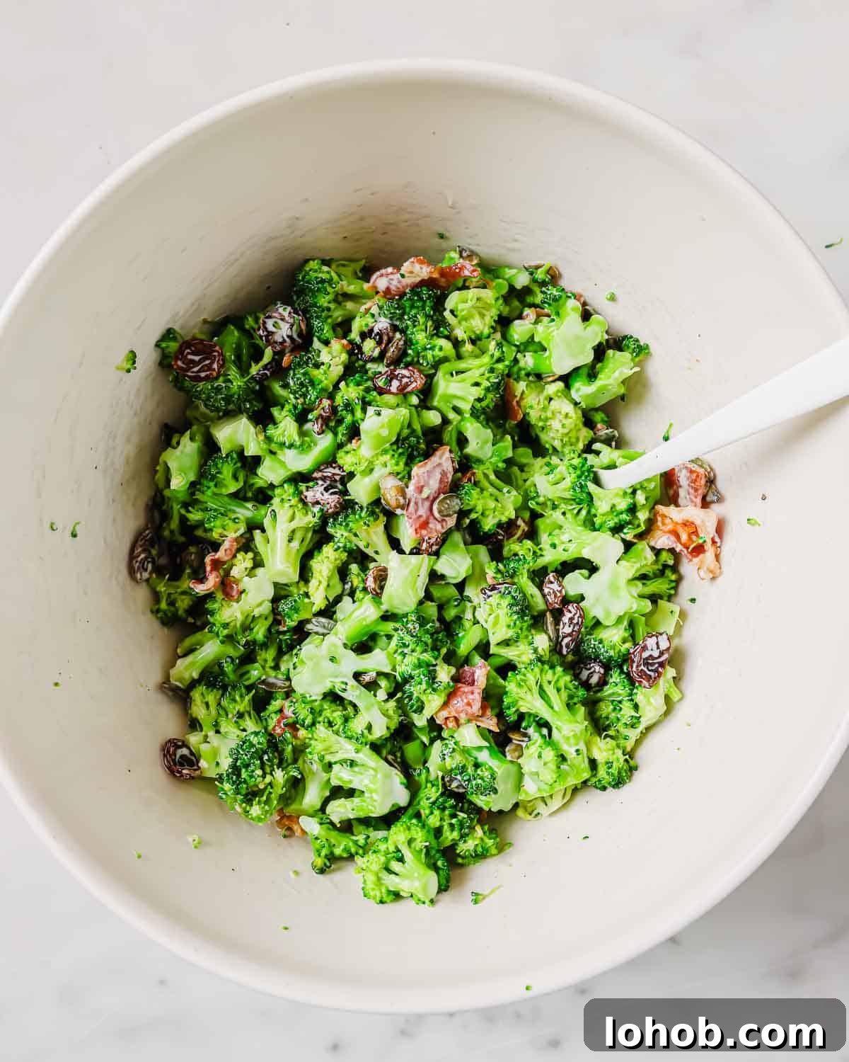 A bowl of broccoli salad with raisins and chopped bacon, mixed in a creamy dressing.