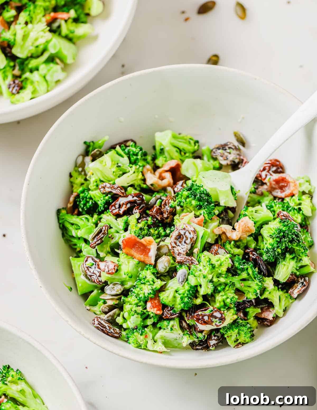 A bowl of broccoli salad with raisins, sunflower seeds, bacon bits, and a creamy dressing, accompanied by a white spoon. Three bowls are partially visible in the background.