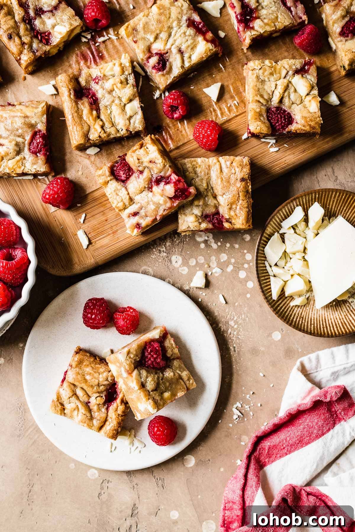 Raspberry White Chocolate Blondies Recipe — Chewy, Fruity Bars 11 Two blondie squares on a small plate with raspberries and more blondie squares nearby.