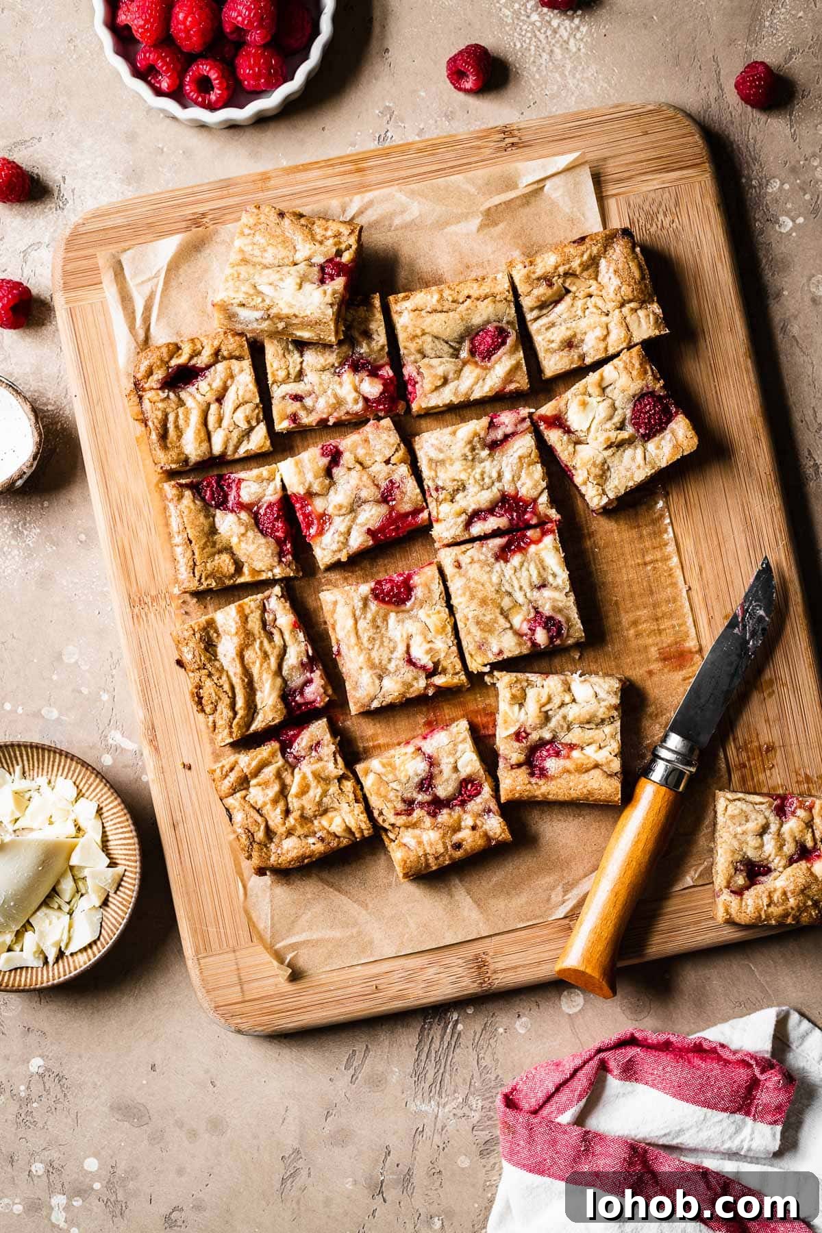 Raspberry White Chocolate Blondies Recipe — Chewy, Fruity Bars 10 Raspberry and white chocolate blondies cut into squares on a wooden cutting board.