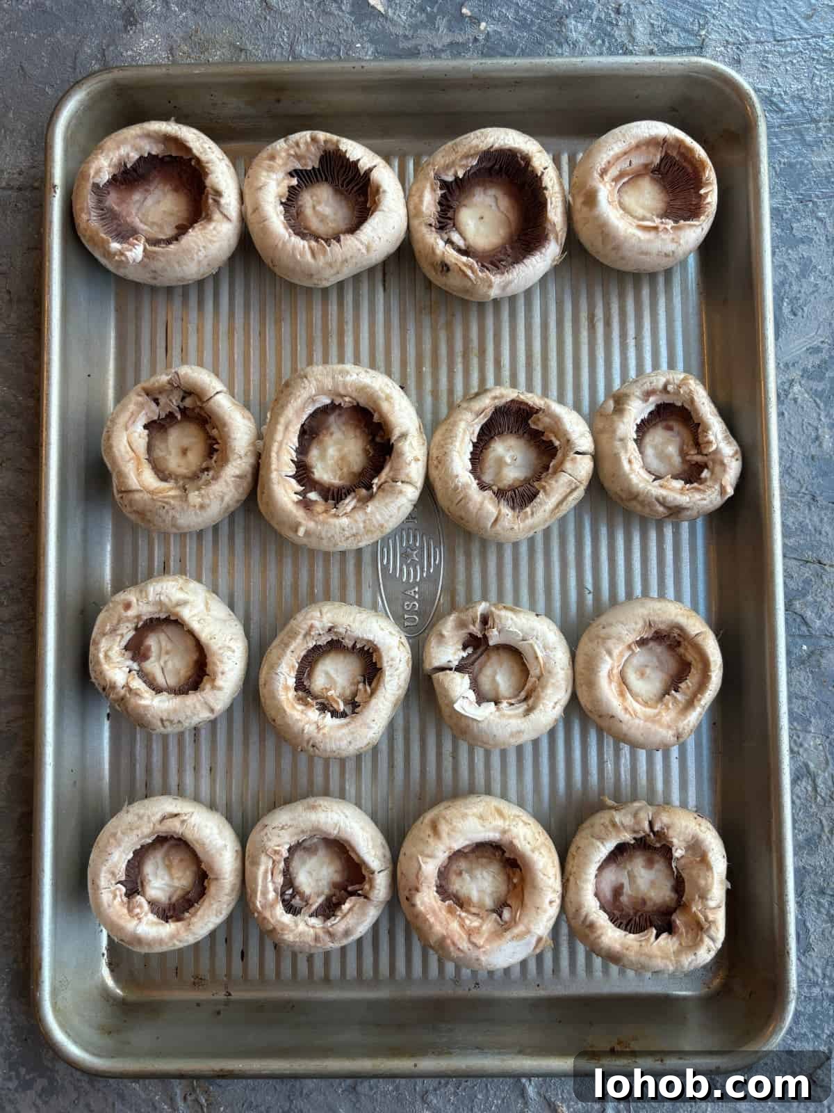 overhead image of a baking sheet topped with white button mushrooms. The mushrooms are clean, the stems have been removed, and they are upside down.
