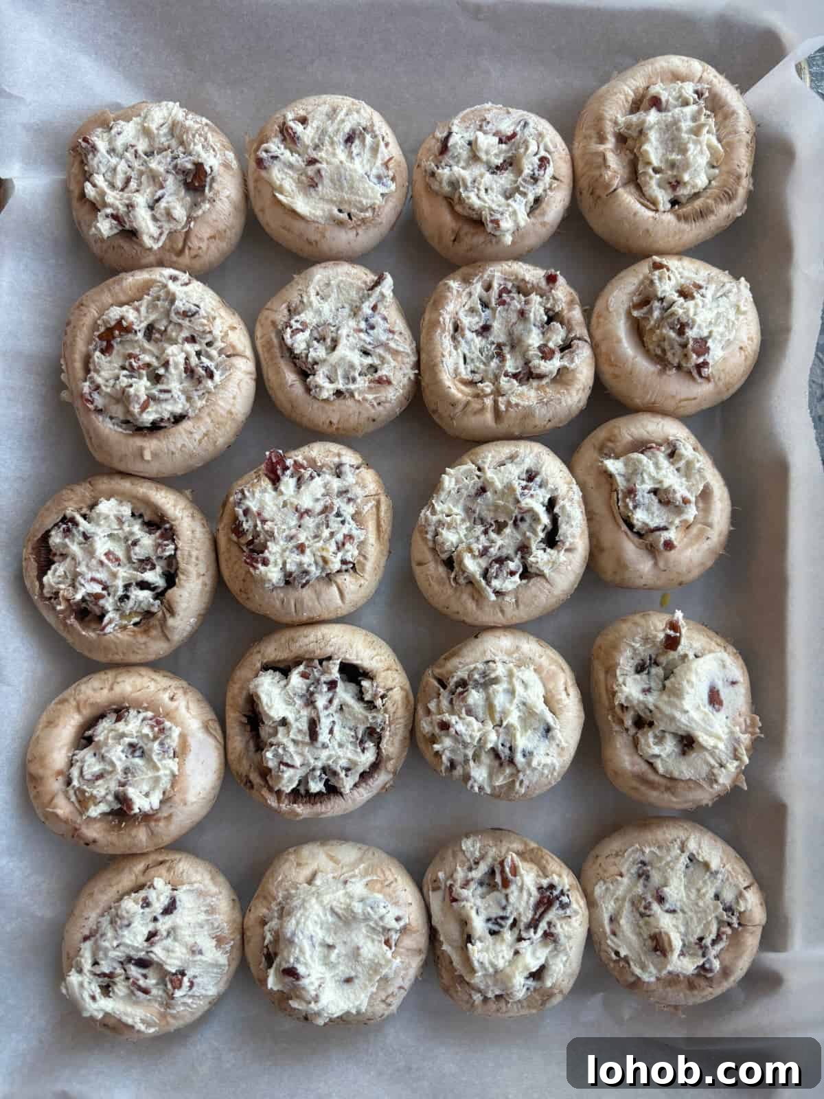 overhead image of a parchment paper lined baking sheet. On top are white button mushrooms that have been filled with a mixture of garlic and herb cheese, chopped pecans, and chopped dried cranberries.