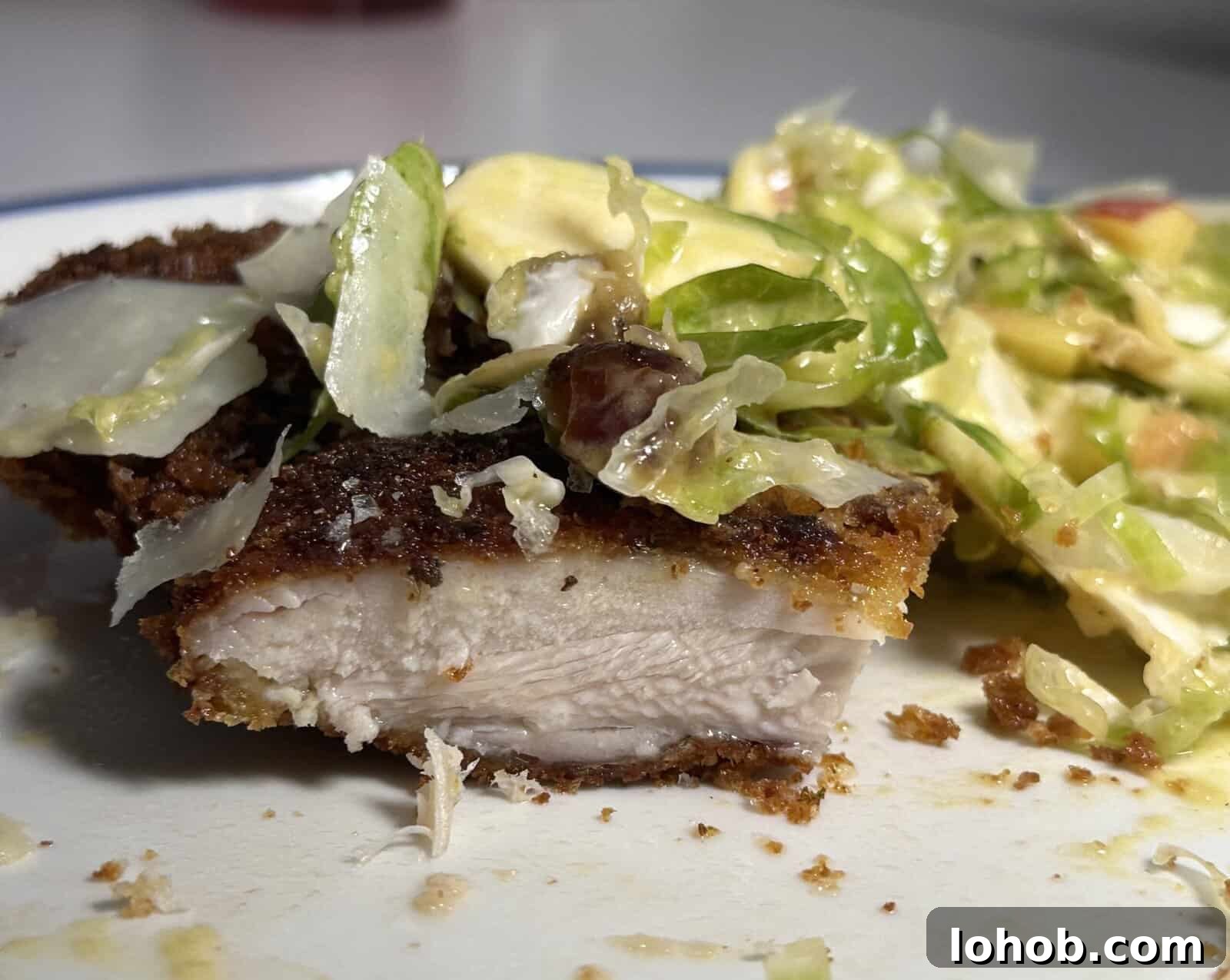 Crispy Chicken Thigh Bites with Shaved Brussels Sprout Salad 3 Close-up of a plate with a piece of breaded, cooked chicken cut in half, showing the white interior, next to a creamy salad with lettuce, avocado, and bacon.