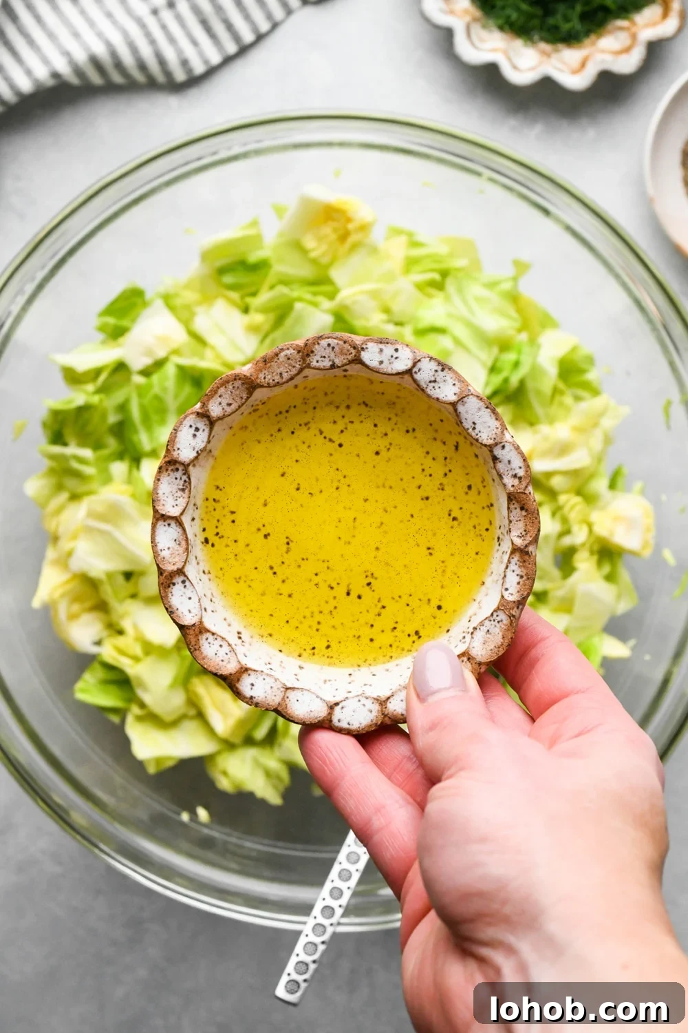 How to make chopped green cabbage salad: A hand adding olive oil from a small ceramic dish to the bowl of cabbage.