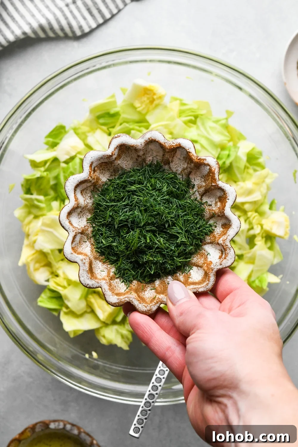 How to make chopped green cabbage salad: A hand adding fresh chopped dill from a small ceramic dish to the bowl of cabbage.