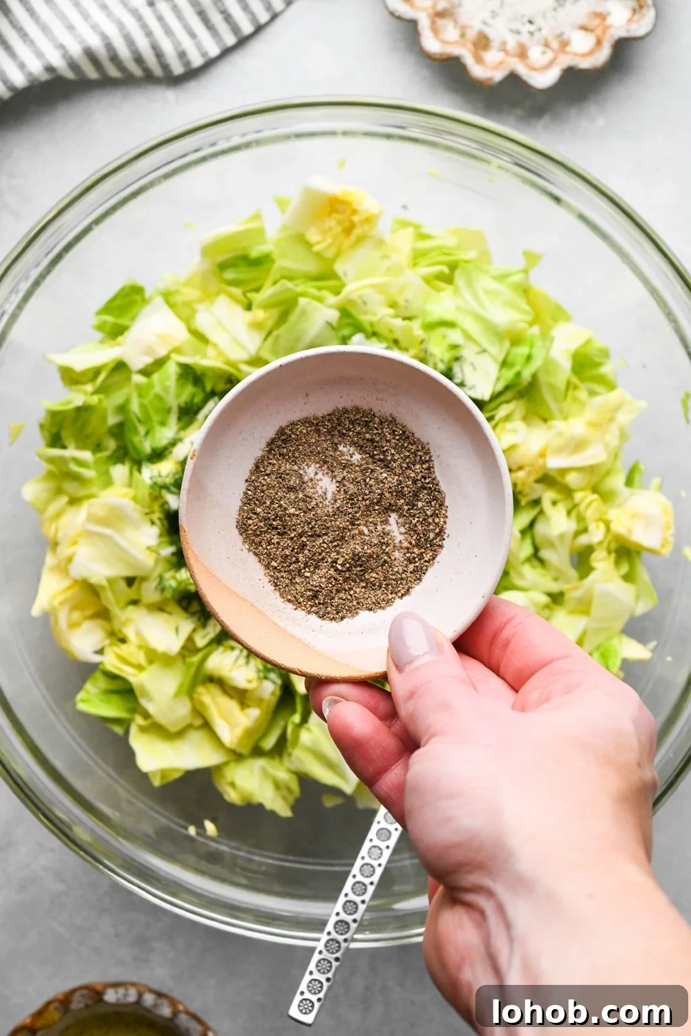 How to make chopped green cabbage salad: A hand adding ground black pepper from a small ceramic dish to the bowl of cabbage.