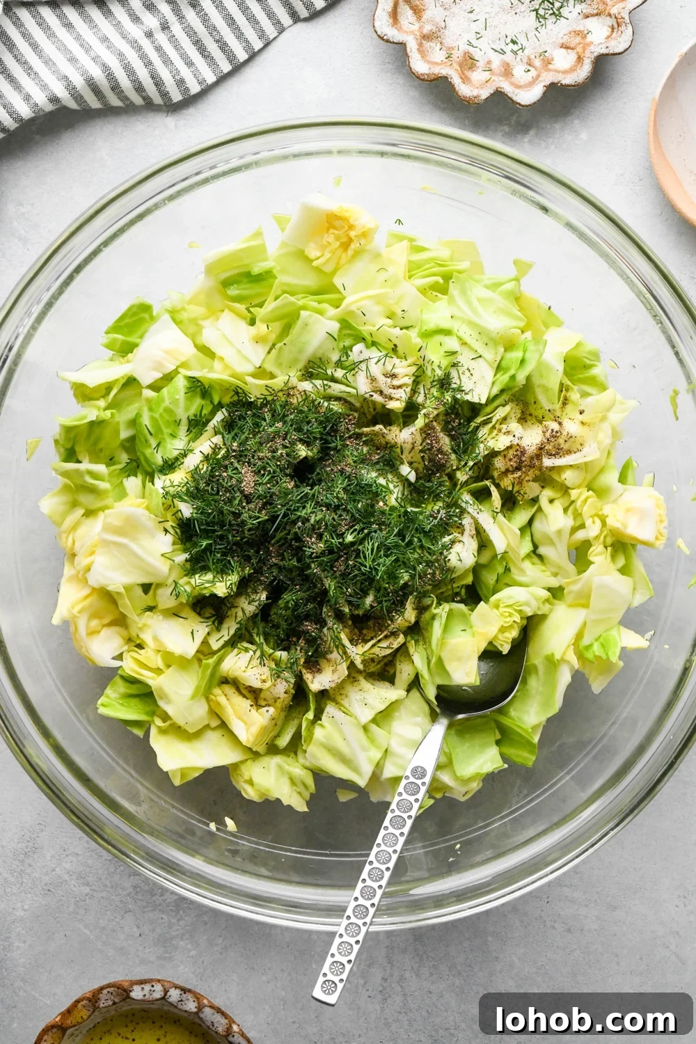 How to make chopped green cabbage salad: Chopped cabbage in glass bowl with olive oil, fresh dill, and black pepper on top before mixing.