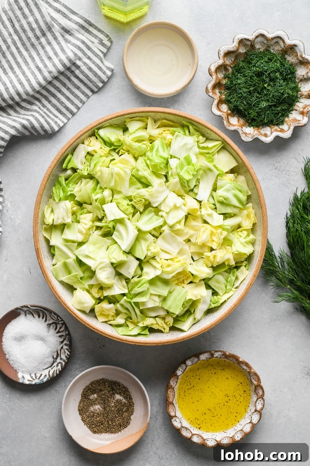 Ingredients for chopped green cabbage salad in various ceramic dishes on a light grey colored background with a striped navy and cream linen peeking into the frame.